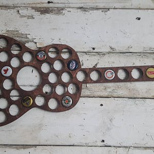 Könnte beinhalten: Ein hölzerner Flaschenverschluss-Halter in Gitarrenform mit brauner Beize. Der Halter hat 40 runde Ausschnitte für Flaschenverschlüsse. Einige der Ausschnitte sind mit Flaschenverschlüssen gefüllt.