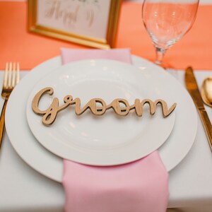 May include: A white plate with a wooden sign that says "groom" on it. The plate is set on a white tablecloth with a pink napkin and gold silverware.