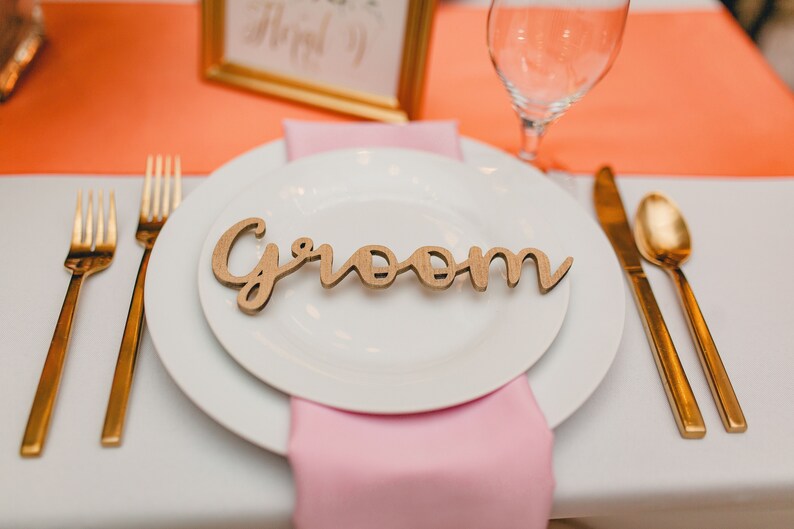 May include: A white plate with a wooden sign that says "groom" on it. The plate is set on a white tablecloth with a pink napkin and gold silverware.