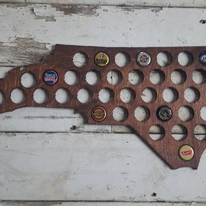 Puede incluir: Una decoración de pared de madera con forma del estado de Carolina del Norte con 49 recortes circulares para tapas de botellas. Los recortes están llenos de varias tapas de botellas, incluidas Miller Lite, Budweiser y Yuengling.