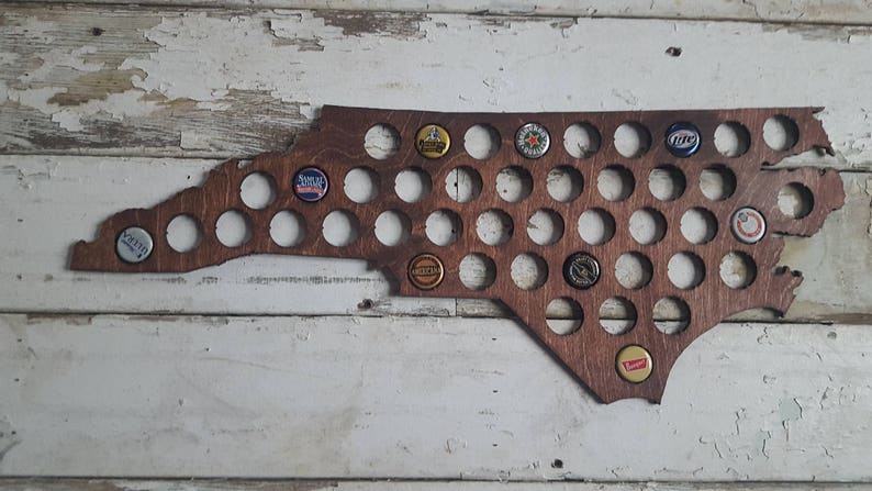 Puede incluir: Una decoraci&oacute;n de pared de madera con forma del estado de Carolina del Norte con 49 recortes circulares para tapas de botellas. Los recortes est&aacute;n llenos de varias tapas de botellas, incluidas Miller Lite, Budweiser y Yuengling.