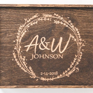 May include: A dark brown wooden box with a hinged lid. The lid is engraved with a floral wreath design and the text "A&W JOHNSON 2-14-2018".