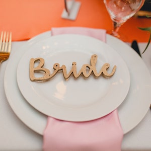 May include: A white plate with a wooden sign that says "Bride" on it. The plate is set on a white tablecloth with a pink napkin and gold silverware.
