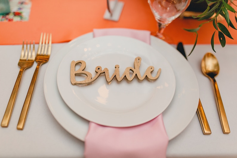 May include: A white plate with a wooden sign that says "Bride" on it. The plate is set on a white tablecloth with a pink napkin and gold silverware.