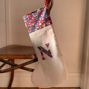 May include: A Christmas stocking made of light beige fabric with a dark blue cuff featuring a red, yellow, and white floral pattern. The letter N is appliqued on the centre of the stocking in the same floral fabric. A red ribbon loop is attached.