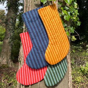 May include: Four quilted fabric stockings hanging together, featuring vertical stripes in distinct colours: bright red, navy blue, golden yellow, and muted sage green. Each stocking has a simple, classic shape with a flat top and a rounded toe.