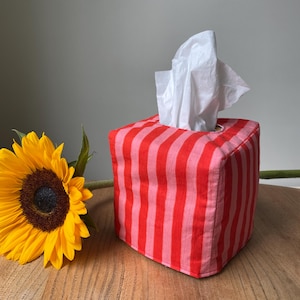 May include: A square fabric tissue box cover featuring vertical stripes in alternating shades of bright red and light pink. A single white tissue is pulled through the top opening. A yellow sunflower with a dark brown centre sits beside it.