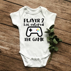 May include: White baby grow with black text that reads "Player 2 has entered the game" and a black and white illustration of a video game controller with coloured buttons.