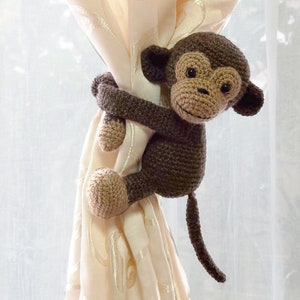 May include: A brown crocheted monkey toy holding a curtain tieback. The monkey has a brown body, tan face, and black eyes.