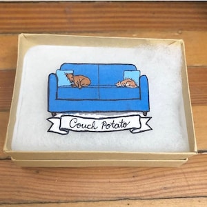 Könnte beinhalten: Eine blaue Couch mit zwei braunen Tieren, die darauf schlafen. Die Couch hat zwei Kissen. Ein Banner unter der Couch sagt "Couch Potato".