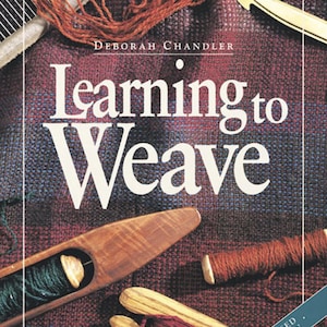 May include: The book Learning to Weave by Deborah Chandler features a cover with a plaid fabric background, a wooden boat shuttle with green yarn, a wooden crochet hook, and two wooden sticks wrapped in red yarn, labeled Revised Edition.