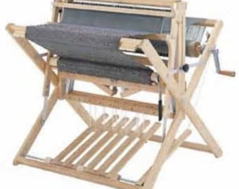 Puede incluir: Un telar de madera con un textil tejido gris en su lugar. El telar tiene un acabado de madera natural y una base en forma de cruz. Una manivela es visible en el lado derecho. El telar está diseñado para crear tejidos.