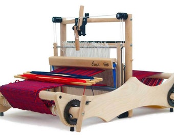 Puede incluir: Un telar de mesa de madera con una tela tejida roja, azul y blanca. El telar tiene una manivela en el lado derecho y una palanca de madera en el lado izquierdo. El telar está etiquetado como "Erica".