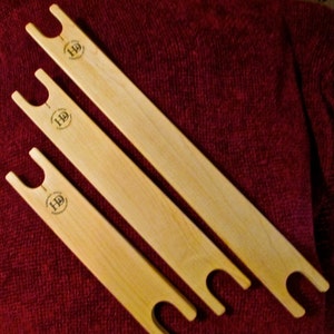May include: Three wooden weaving tools with a rounded handle and two loops at the end. Each tool has the letters "HD" printed on the surface.