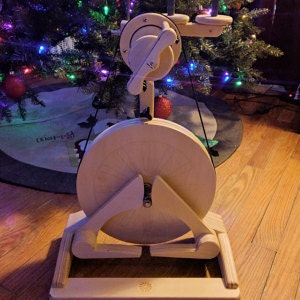 May include: A wooden spinning wheel with a large, round wheel and a smaller, round wheel with a handle. The spinning wheel is made of light wood and has a natural finish. The spinning wheel is sitting on a wooden floor.