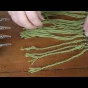 May include: Close-up of green yarn with fringe being tied off with a knot. The yarn is being held in place with silver clips.