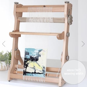 May include: A wooden weaving loom with a 16-inch weaving width. The loom has a natural wood finish and features a partially woven tapestry with blue, yellow, and black colors. The loom is designed for creating woven textiles.