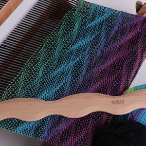 May include: A wooden weaving shuttle with a curved handle and a hook on one end. The shuttle is lying on top of a loom with a colorful woven fabric. The fabric is a gradient of blue, green, purple, and black. The text "Ashford" is printed on the shuttle.