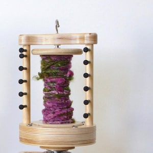 May include: A wooden yarn swift with a pink and green yarn ball wound on it. The swift has a crank handle and a hook on top.