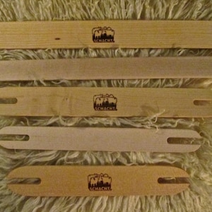 May include: Four wooden weaving shuttles with a logo that says "Schacht" on each. The shuttles are arranged in a stack, with the largest shuttle on the bottom and the smallest shuttle on top.