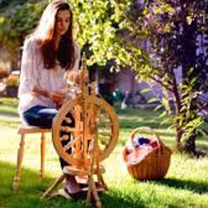 May include: A woman sits on a wooden stool in a grassy yard, spinning yarn on a wooden spinning wheel. A basket with yarn balls sits next to the wheel.