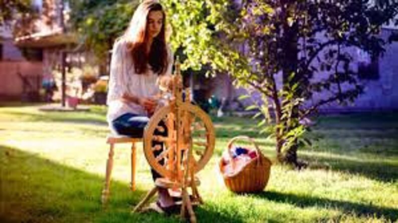 May include: A woman sits on a wooden stool in a grassy yard, spinning yarn on a wooden spinning wheel. A basket with yarn balls sits next to the wheel.