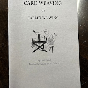 May include: A black and white illustration of a person weaving on a loom. The book cover reads "CARD WEAVING OR TABLET WEAVING" and "By Russell E. Groff Distributed by Unicorn Books and Crafts, Inc."