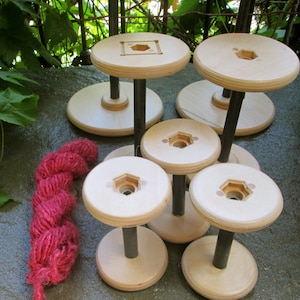 May include: Six wooden yarn swift spools with a red ball of yarn on a gray stone surface. The spools are stacked and have a black metal center post.