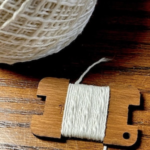 May include: A wooden yarn swift with white yarn wrapped around it, next to a large ball of white yarn. The swift is a light brown color and has a small hole on one end. The yarn is a soft, off-white color.