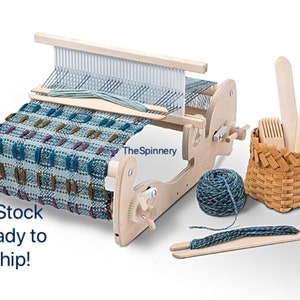 May include: A wooden tabletop loom with a partially woven tapestry in shades of blue, green, brown, and purple. The loom includes a wooden shuttle, a ball of yarn, a small basket with weaving tools, and a wooden dowel with yarn attached. The text "The Spinnery" is visible on the loom.