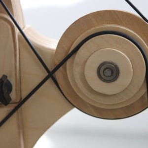 May include: A wooden pulley system with a black rubber belt. The pulley is made of multiple layers of wood and has a metal bearing in the center.
