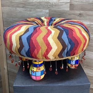 May include: A round, upholstered ottoman with a colorful chevron pattern in red, blue, yellow, and brown. The ottoman has three decorative, painted legs and beaded fringe. The ottoman is approximately 18 inches in diameter.