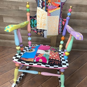May include: A rocking chair featuring a patchwork fabric backrest and seat with various colorful patterns. The frame consists of multi-colored spindles in yellow, pink, blue, and green with gold accents. The seat edge has a black and white checkerboard pattern.