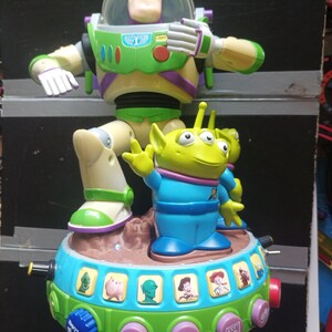 May include: A colorful Toy Story-themed toy featuring Buzz Lightyear and an alien figure. The toy has a green and blue base with character images and buttons. Buzz Lightyear is in a green and white spacesuit. The alien is green with three eyes.