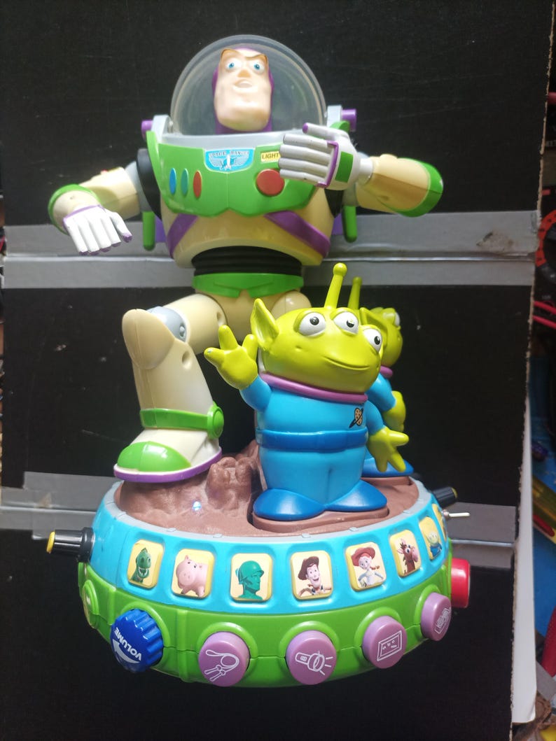 May include: A colorful Toy Story-themed toy featuring Buzz Lightyear and an alien figure. The toy has a green and blue base with character images and buttons. Buzz Lightyear is in a green and white spacesuit. The alien is green with three eyes.