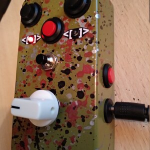 May include: A green and black speckled effect guitar pedal with a white knob, two black knobs, a red button, and a black foot switch. The pedal has a power input and a red LED light.