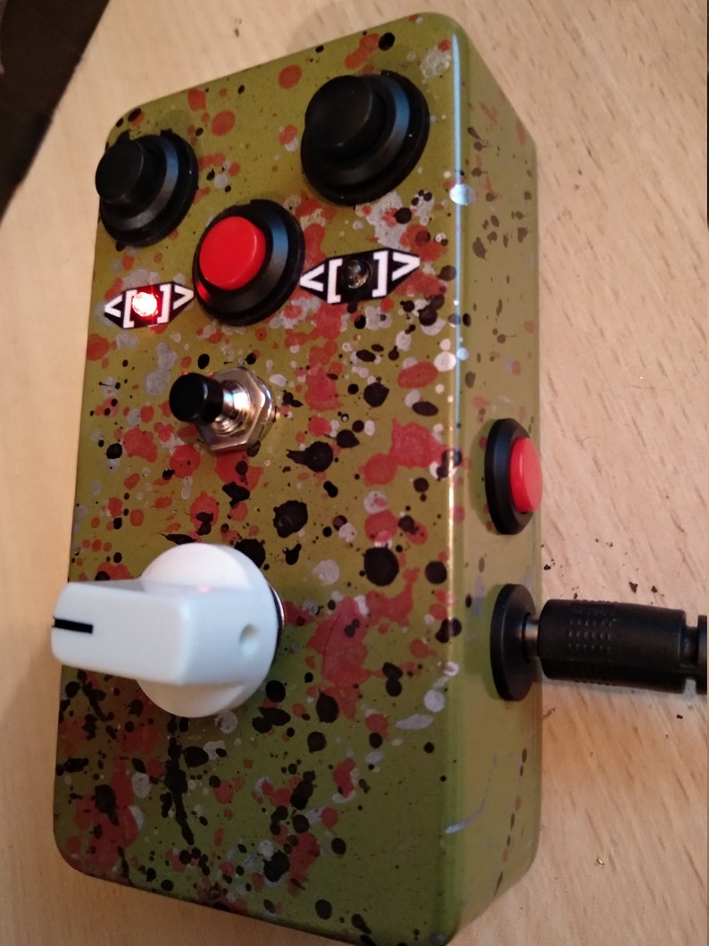 May include: A green and black speckled effect guitar pedal with a white knob, two black knobs, a red button, and a black foot switch. The pedal has a power input and a red LED light.