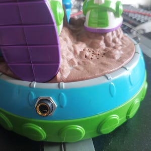 May include: A colorful toy featuring a round base with a blue and green exterior. The top has a brown, textured surface with small holes. A purple and white element is visible, along with other green and white details.