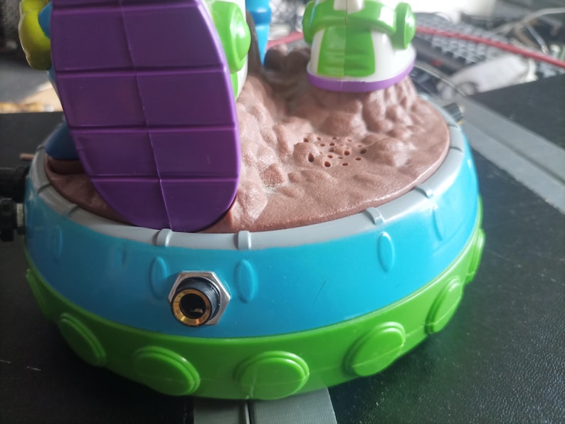 May include: A colorful toy featuring a round base with a blue and green exterior. The top has a brown, textured surface with small holes. A purple and white element is visible, along with other green and white details.