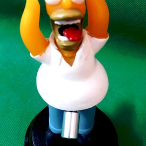 May include: A Homer Simpson figurine with arms raised, mouth open, and tongue sticking out. The figure has a yellow head, orange arms, a white shirt, blue pants, and is mounted on a black base with an orange button.