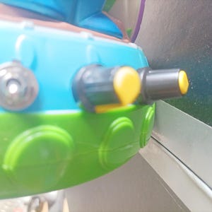 May include: Close-up of a toy vehicle with a blue top, green base, and yellow accents. Features include a black and yellow nozzle and a silver-colored circular detail. The toy is designed with rounded shapes and bright colors.