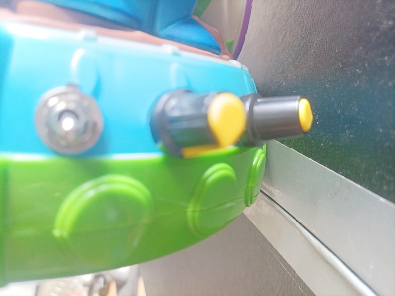 May include: Close-up of a toy vehicle with a blue top, green base, and yellow accents. Features include a black and yellow nozzle and a silver-colored circular detail. The toy is designed with rounded shapes and bright colors.