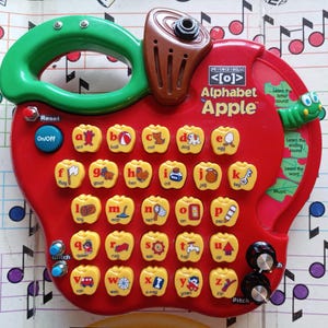 May include: A red, apple-shaped educational toy with a green handle and brown speaker. The toy features yellow buttons with letters and corresponding images, labeled "Alphabet Apple". Includes buttons for sound and music.
