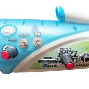 May include: A futuristic-looking toy instrument in shades of blue, white, and yellow. It features knobs, switches, and the text "Glitch Buzz Drive Ranger." The design includes a meter and a small, white, cylindrical component.
