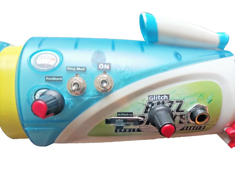 May include: A futuristic-looking toy instrument in shades of blue, white, and yellow. It features knobs, switches, and the text "Glitch Buzz Drive Ranger." The design includes a meter and a small, white, cylindrical component.