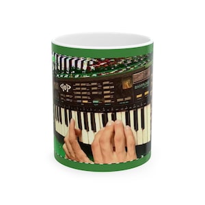 May include: A white ceramic mug with a green rim and handle. The mug features a graphic of a vintage synthesizer keyboard with hands playing the keys. The mug's design includes the text "<0>".