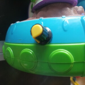 May include: Close-up of a colorful toy with a blue and green base. A yellow knob with a black center is visible on the side. The toy has a brown and purple top.