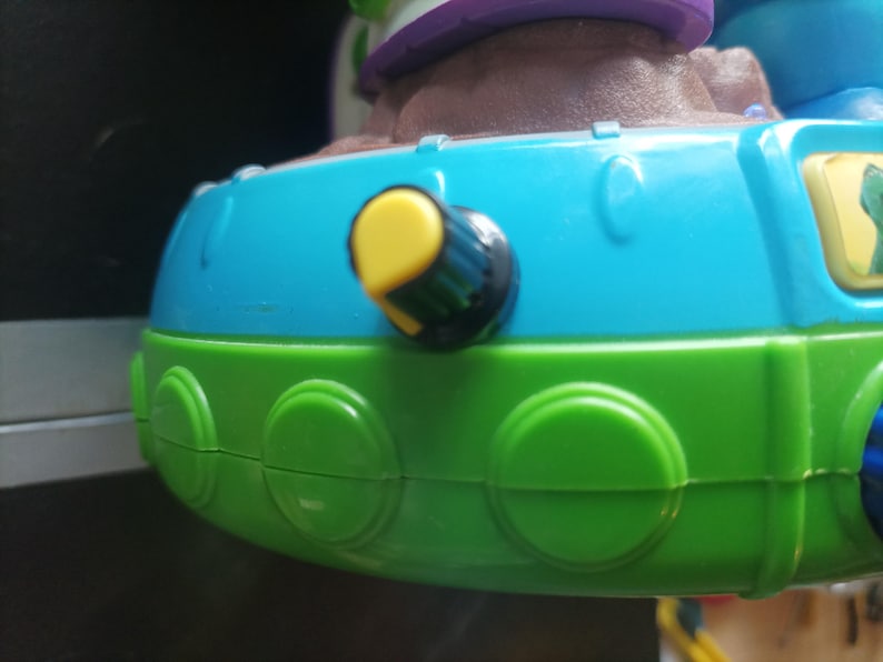 May include: Close-up of a colorful toy with a blue and green base. A yellow knob with a black center is visible on the side. The toy has a brown and purple top.