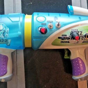 May include: A colorful toy water gun with a blue, white, yellow, and purple design. The toy has the word "Buzz" printed on the side and a red dial. It has a trigger and a handle.
