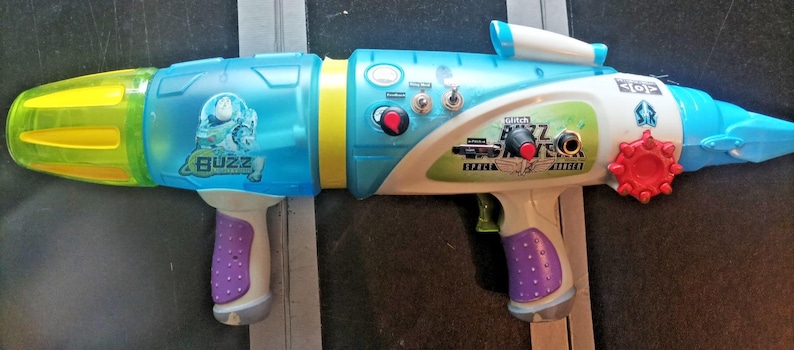 May include: A colorful toy water gun with a blue, white, yellow, and purple design. The toy has the word "Buzz" printed on the side and a red dial. It has a trigger and a handle.